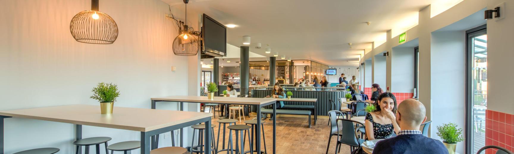 Photo of a cafe seating area with high rectangular tables surrounded by stools and round tables with chairs