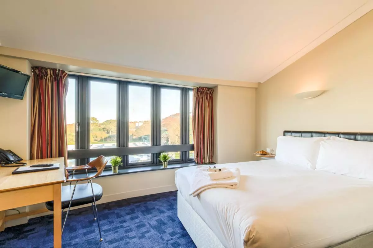 Photo of a hotel room with large double bed, desk, chair and large windows